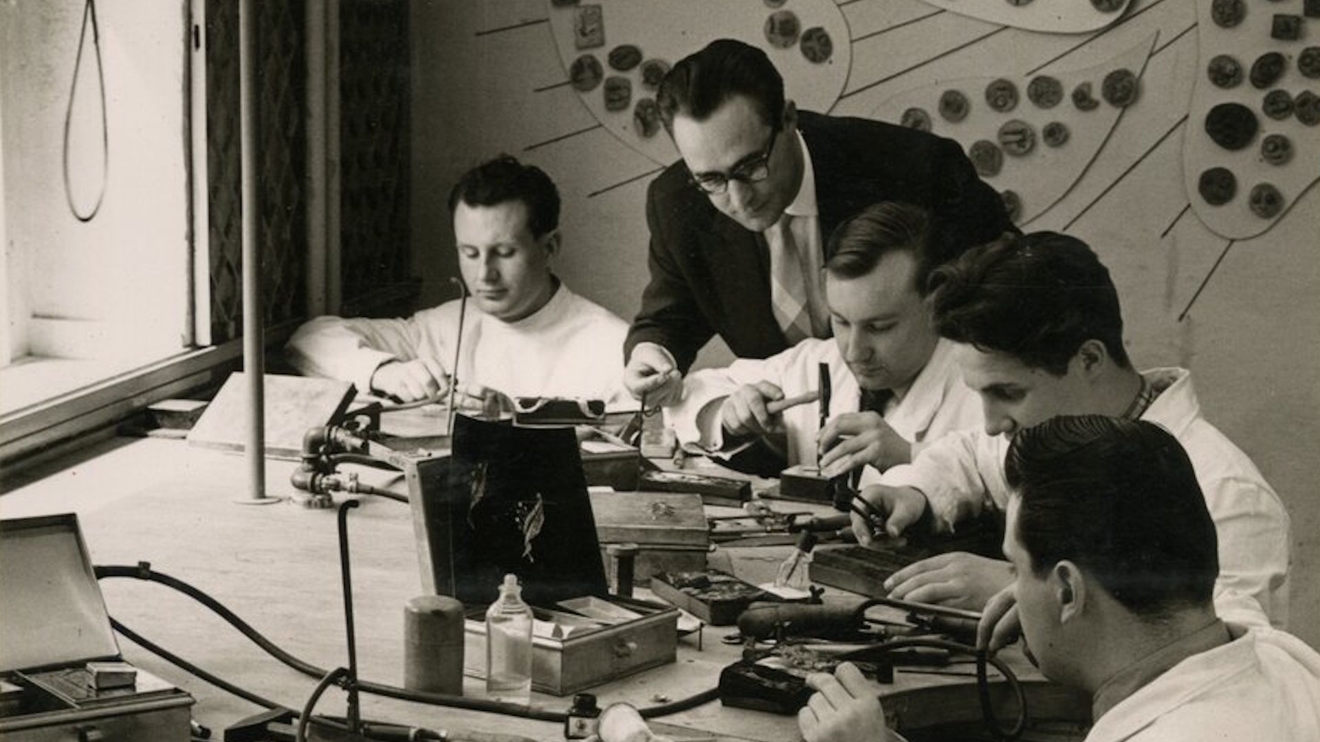 René Kern steht Ende der 1950er Jahre in seinem Düsseldorfer Atelier hinter mehreren Goldschmieden bei der Schmuckfertigung.